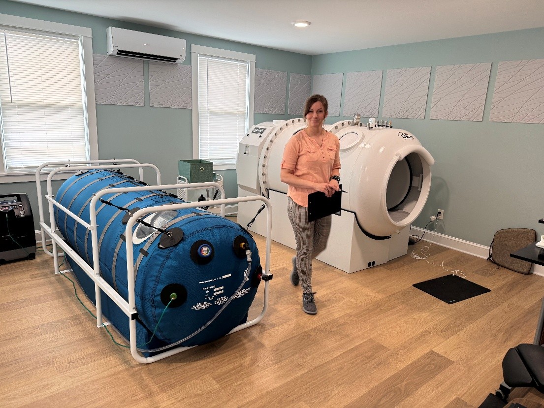 A wellness professional standing between a blue portable soft-shell hyperbaric chamber and a white hard-shell HBOT chamber in a professional treatment room.